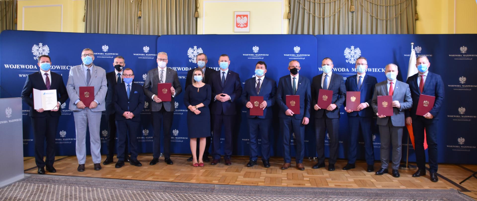 Na zdjęciu znajduje się czternaście elegancko ubranych osób - trzynastu mężczyzn i jedna kobieta. Osoby te pozują na granatowej ściance wizerunkowej Wojewody Mazowieckiego. Większość z nich trzyma w rękach bordowe teczki ze złotym orłem - Godłem RP. W teczkach tych znajdują się podpisane wcześniej umowy. 