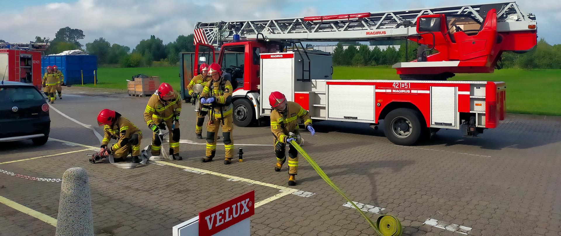 Na zdjęciu widać jak strażacy rozwijają węże strażackie. W tle widać drabinę mechaniczną. Pogoda słoneczna