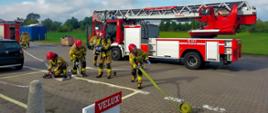 Na zdjęciu widać jak strażacy rozwijają węże strażackie. W tle widać drabinę mechaniczną. Pogoda słoneczna