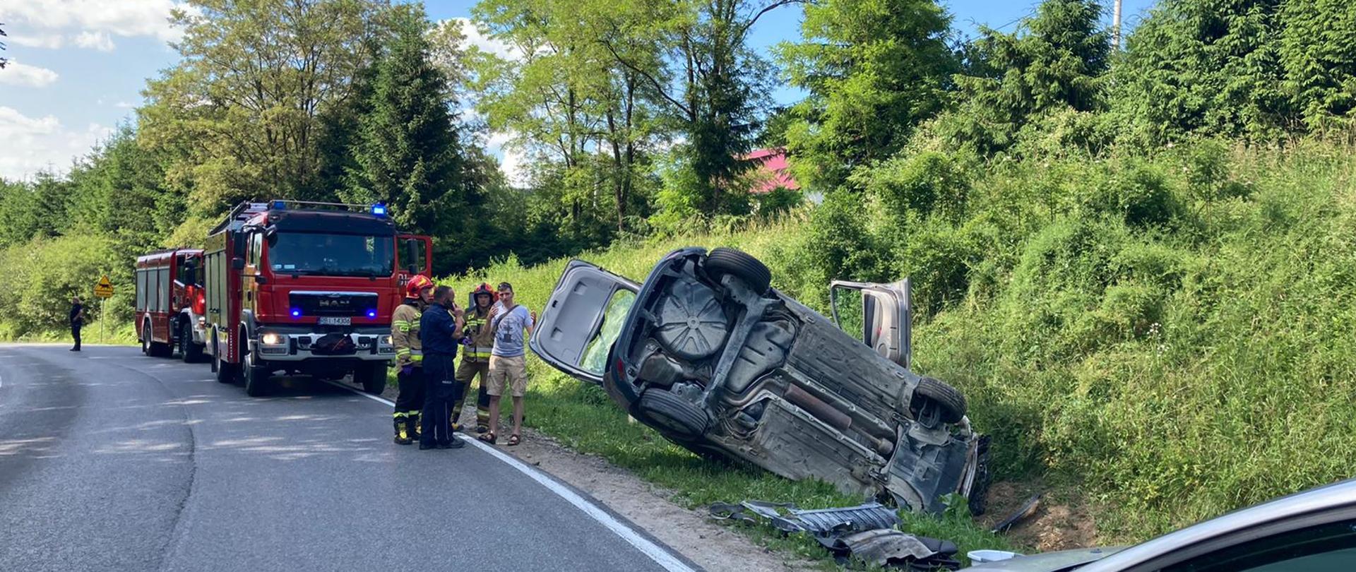 Na zdjęciu widać ustawione przy drodze na zakręcie dwa samochody pożarnicze, dwóch strażaków PSP, policjanta oraz osobę postronną. Obok nich znajduje się samochód osobowy w rowie który leży na boku, jest po wypadku. W prawym dolnym rogu zdjęcia widać przód innego samochodu osobowego. Krajobraz to drzewa, trawa i czerwony dach domu w oddali.