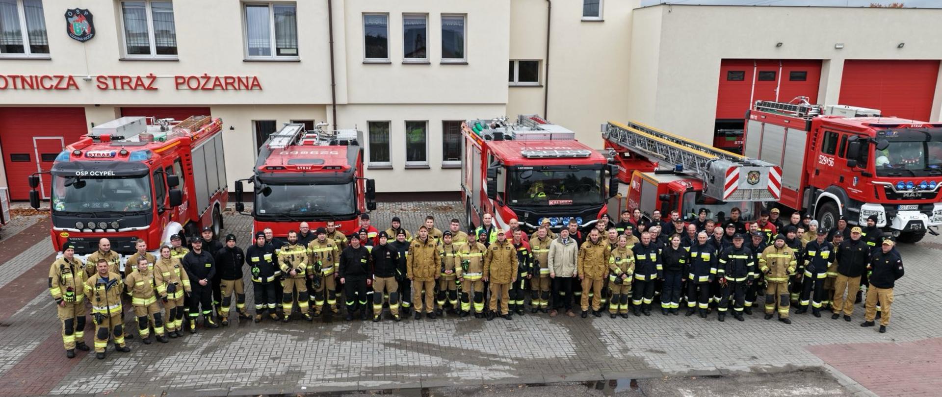 Zdjęcie przedstawia wszystkich uczestników ćwiczeń, ustawieni w półksięzycu na tle remizy osp i samochodów pożarniczych, strażacy w mundurach, na budynku napis Ochotnicza Straż Pożarna.