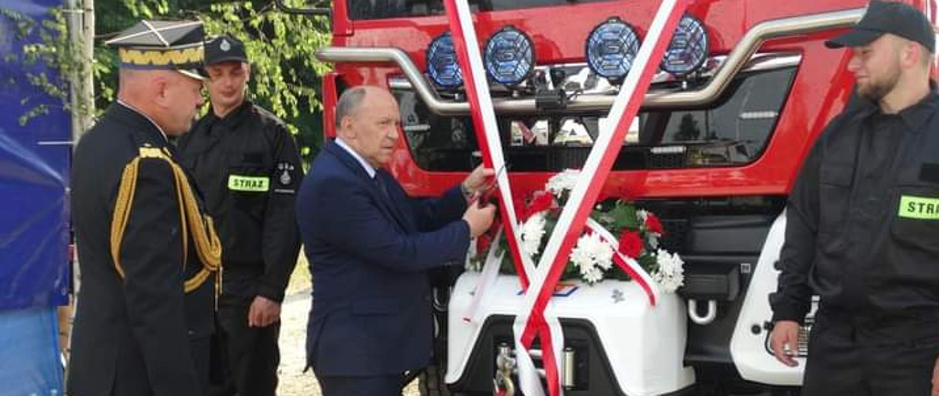 Na zdjęciu strażak PSP w umundurowaniu galowym, dwóch strażaków w ubraniach koszarowych oraz jedna osoba cywilna, która przecina biało czerwoną wstęgę zawieszoną na przodzie samochodu strażackiego.