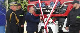 Na zdjęciu strażak PSP w umundurowaniu galowym, dwóch strażaków w ubraniach koszarowych oraz jedna osoba cywilna, która przecina biało czerwoną wstęgę zawieszoną na przodzie samochodu strażackiego.