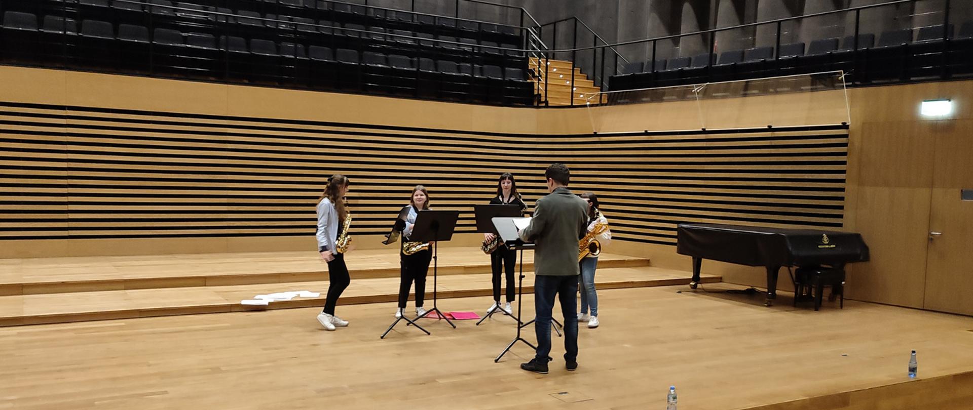 Mężczyzna stoi przy pulpicie na scenie sali koncertowej PSM, tyłem do widowni, przed nim również przy pulpitach stoją cztery uczennice trzymające w rękach saksofony, przodem do widowni. Po lewej stronie leży otwarty futerał na saksofon.