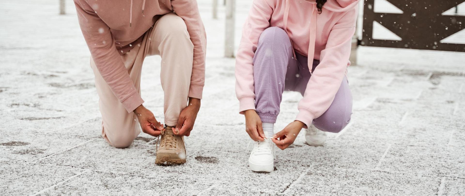 Na zdjęciu jest kobieta i mężczyzna zawiązujący buty w pozycji klęczącej. W tle widać śnieg.