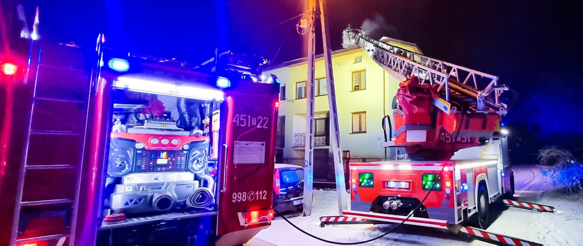 Na pierwszym planie stoją dwa samochody strażackie, w tym jeden wóz drabina przed budynkiem gdzie palą się sadze w kominie. Drabina mechaniczna jest wysunięta przed komin. W koszu drabiny stoi strażak, który gasi pożar sadzy. Aura zimowa, nocna.