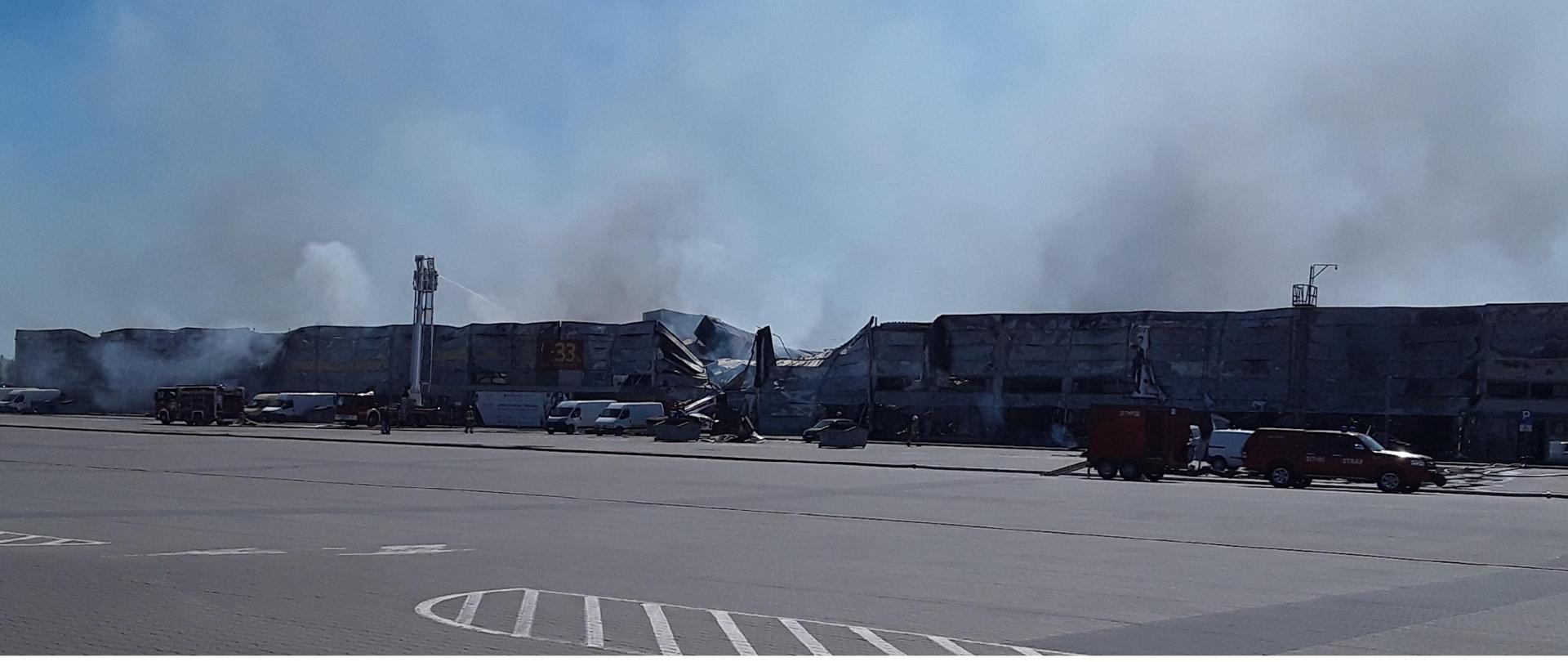 Płonąca hala znajduje się za pustym, asfaltowym parkingiem. Z budynku wydobywa się dym. Środkowa cześć hali jest zawalona.