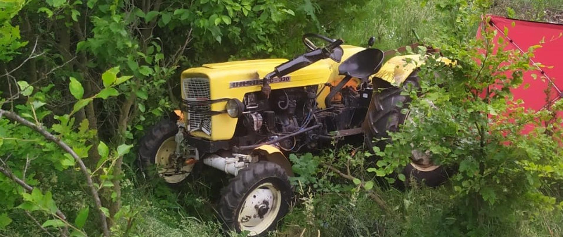 Na zdjęciu widzimy ciągnik rolniczy z uszkodzonymi elementami nadwozia i silnika. Z prawej strony częściowo widoczny parawan osłaniający osobę poszkodowaną. Na drugim planie stoi samochód strażacki. Tło zdjęcia stanowią zarośla i drzewa.