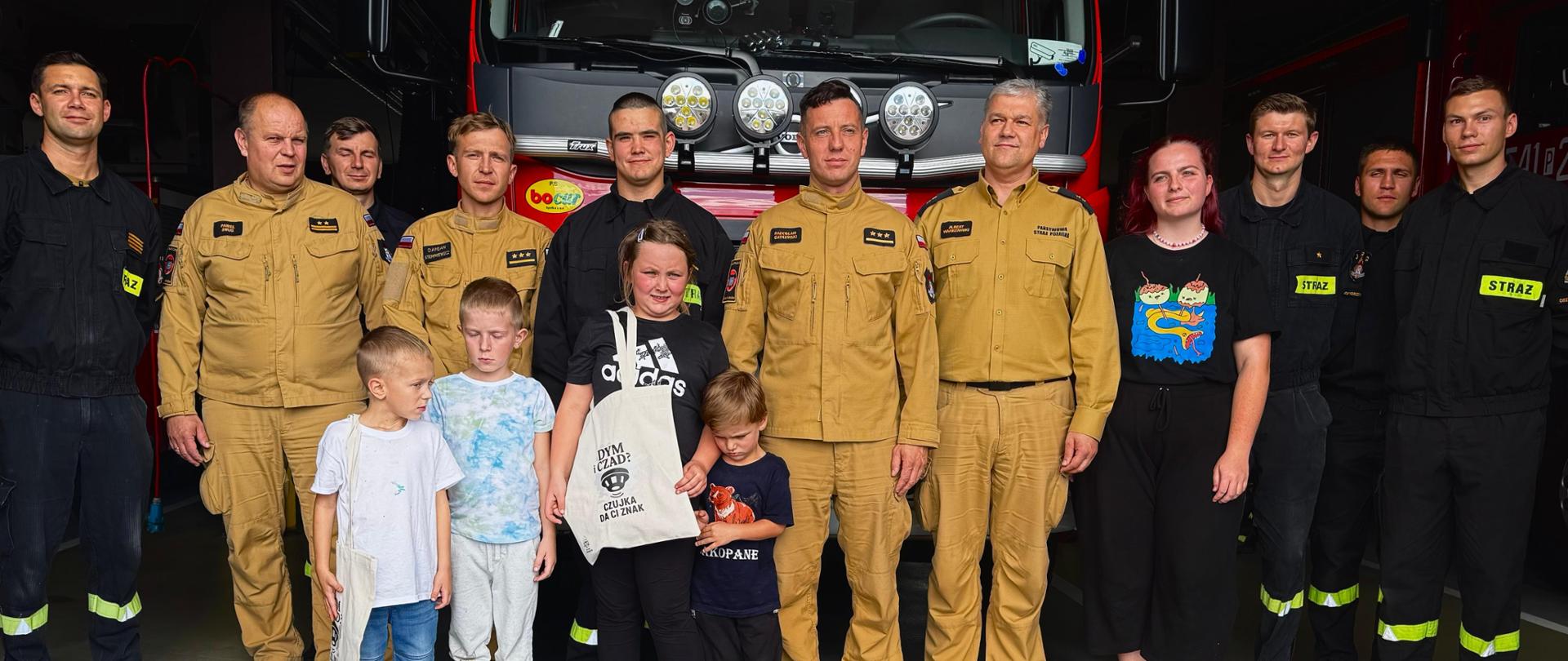 Strażacy wspólnie z uczestnikami wycieczki pozują do zdjęcia przed wozem strażackim. Zdjęcie wykonane w garażu.