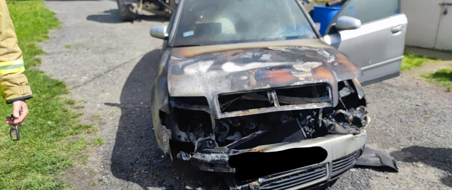 Dzień . Widok na samochód osobowy ze spaloną komorą silnika . Maska pojazdu zamknięta - cały lakier uległ spaleniu. Z przodu pojazdu brak elementów plastikowych ( zderzak, grill, lampy), które uległy spaleniu. Pod maską widać spalone elementy silnika. Drzwi od strony kierowcy otwarte. 