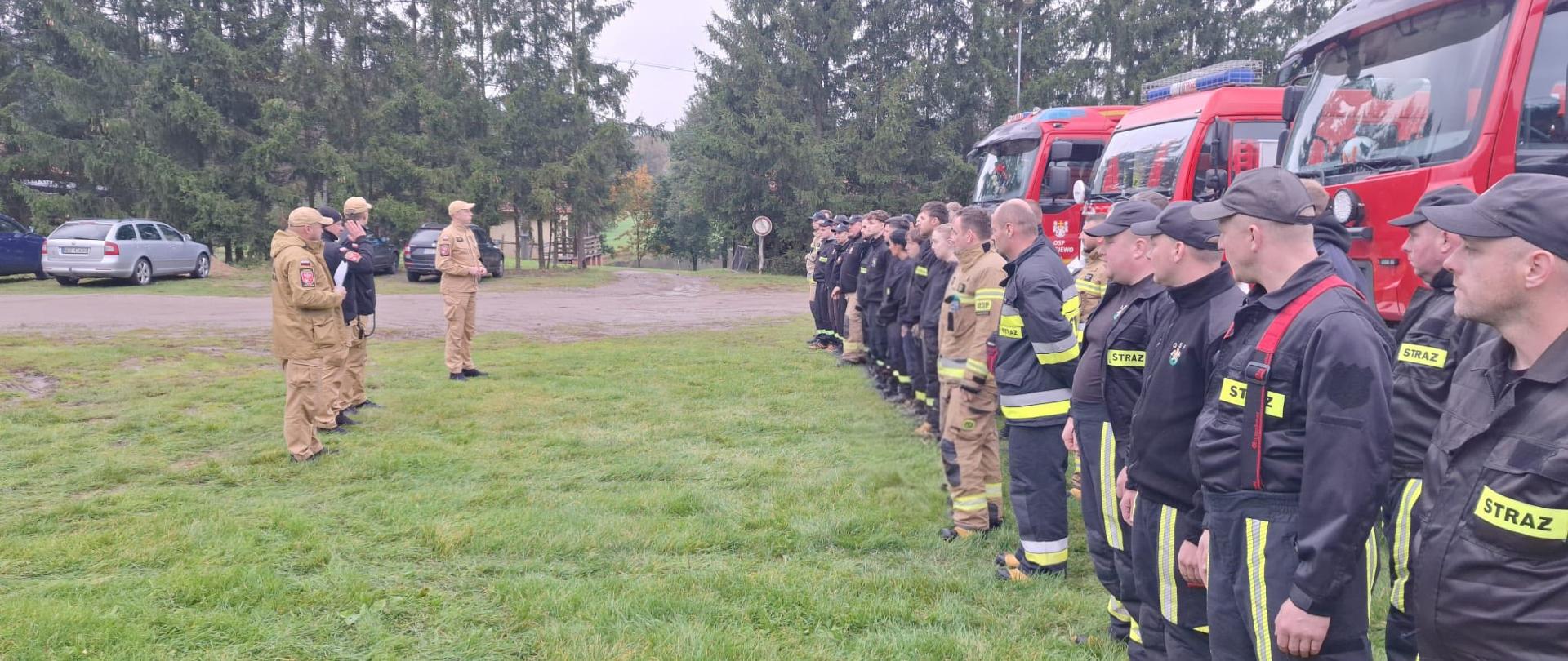 Druhowie ochotniczych straży pożarnych stoją na polanie w szeregu. Maja na sobie mundury. Za nimi, po prawej stronie stoją pojazdy pożarnicze. Przed nimi stoi czterech funkcjonariuszy państwowej Straży Pożarnej.