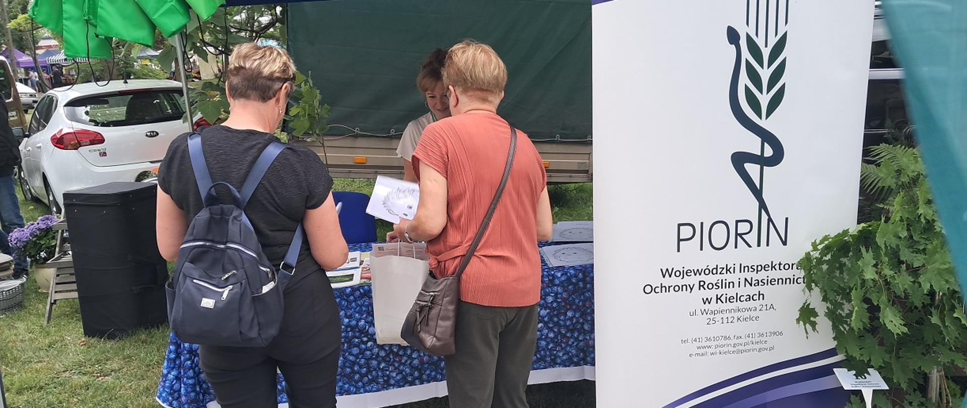 Na zdjęciu widoczne trzy kobiety, dwie zwrócone plecami do zdjęcia. Po prawej stronie roll up z logo PIORiN. Z tyłu po lewej stoi samochód. 