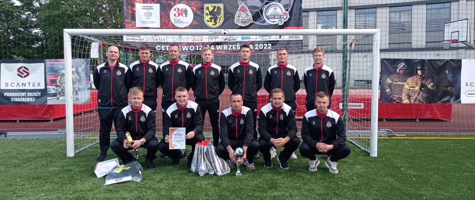 Grupowe zdjęcie drużyny strażaków PSP reprezentujących województwo opolskiego. Strażacy w strojach sportowych na tle bramki.