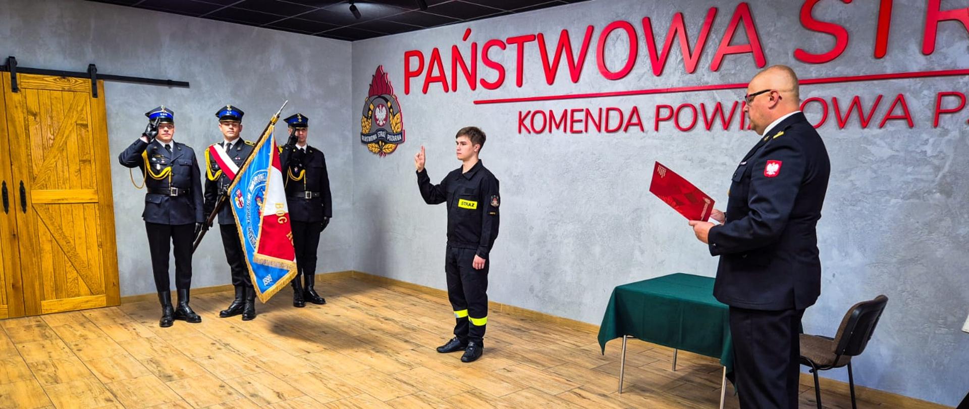 Strażak składa ślubowanie, które odczytuje komendant. W tle stoi poczet sztandarowy.