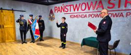 Strażak składa ślubowanie, które odczytuje komendant. W tle stoi poczet sztandarowy.