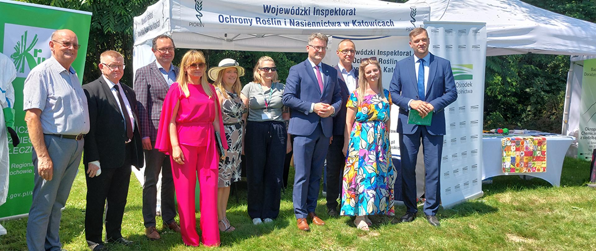 **Audiodeskrypcja zdjęcia:**
Na zdjęciu przedstawiono grupę dziesięciu osób pozujących do wspólnego zdjęcia podczas wydarzenia plenerowego. Uczestnicy stoją na trawie przed białym namiotem wystawowym z napisem „Wojewódzki Inspektorat Ochrony Roślin i Nasiennictwa w Katowicach”. W tle widoczne są drzewa, co wskazuje na park lub teren zielony.
Od lewej strony stoi mężczyzna w jasnoniebieskiej koszuli z krótkim rękawem i jasnych spodniach. Obok niego znajduje się starszy mężczyzna w czarnym garniturze, białej koszuli i krawacie. Kolejna osoba to mężczyzna w marynarce w kratę, jasnej koszuli i okularach.
W centralnej części zdjęcia wyróżnia się kobieta ubrana w intensywnie różowy kombinezon z długimi, szerokimi rękawami. Obok niej stoi kobieta w jasnym kapeluszu i sukience w czarno-biały roślinny wzór. Następnie widoczna jest kobieta w okularach przeciwsłonecznych, szarej bluzce i granatowych spodniach.
Po prawej stronie centrum stoi mężczyzna w granatowym garniturze, białej koszuli i różowym krawacie. Obok niego znajduje się mężczyzna w jasnej koszuli i okularach. Dalej stoi uśmiechnięta kobieta w długiej, kolorowej sukience z abstrakcyjnym wzorem w odcieniach niebieskiego, żółtego, różowego i białego. Skrajnie po prawej widoczny jest wysoki mężczyzna w granatowym garniturze i niebieskim krawacie, trzymający zieloną teczkę.
Po obu stronach namiotu ustawiono roll-upy i stoiska promocyjne związane z instytucjami rolniczymi. Po prawej stronie znajduje się stolik przykryty białym obrusem, na którym leżą materiały informacyjne oraz kolorowa tkanina dekoracyjna. Zdjęcie wykonano w słoneczny dzień, a wszyscy uczestnicy zwróceni są w stronę fotografa i pozują do pamiątkowej fotografii.
