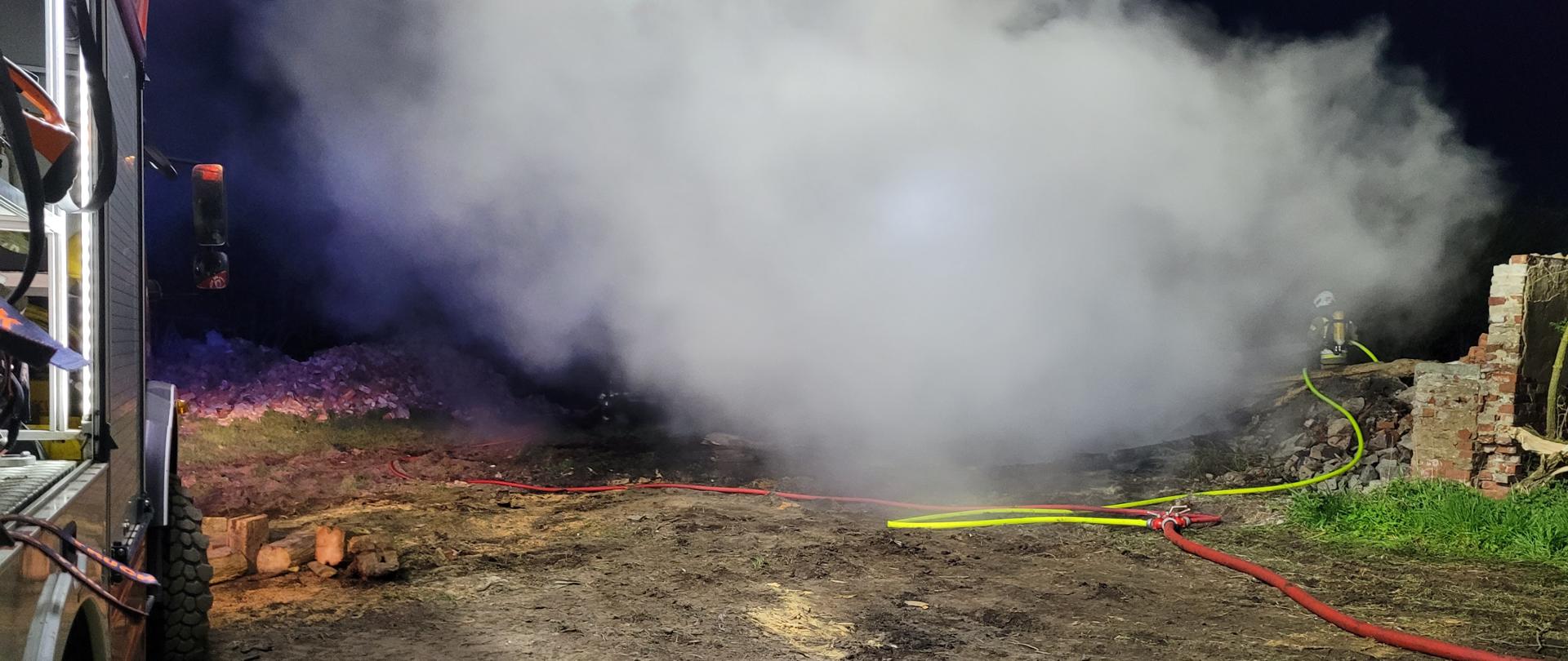 Po lewej stronie fotografii znajduje się wóz strażacki. W centrum zdjęcia palą się zgliszcza budynku. W dymie widać strażaków gaszących pożar. Cały teren oświetlony jest sztucznym światłem. Zdjęcie wykonano w porze nocnej.