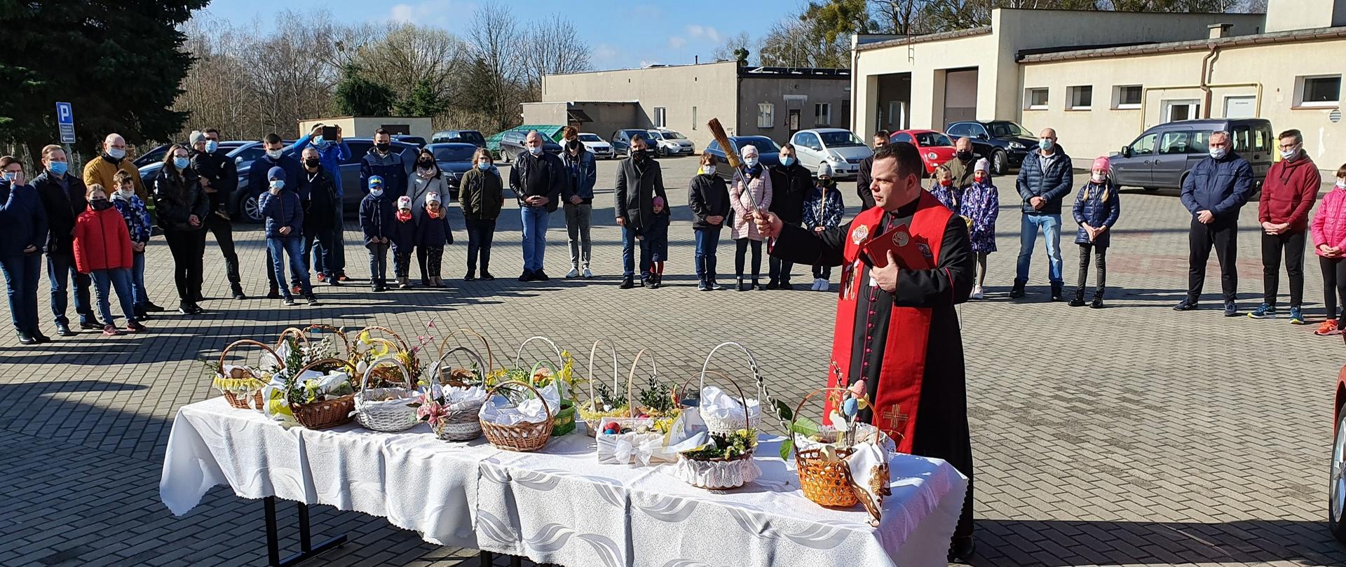 Zdjęcie przedstawia księdza kanonika Mariusza Stasiaka kapelana strażaków w województwie kujawsko-pomorskim podczas poświęcenia pokarmów. Na pierwszym planie stoły z przyniesionymi koszyczkami z pokarmem. W oddali pracownicy Komendy wraz z rodzinami.