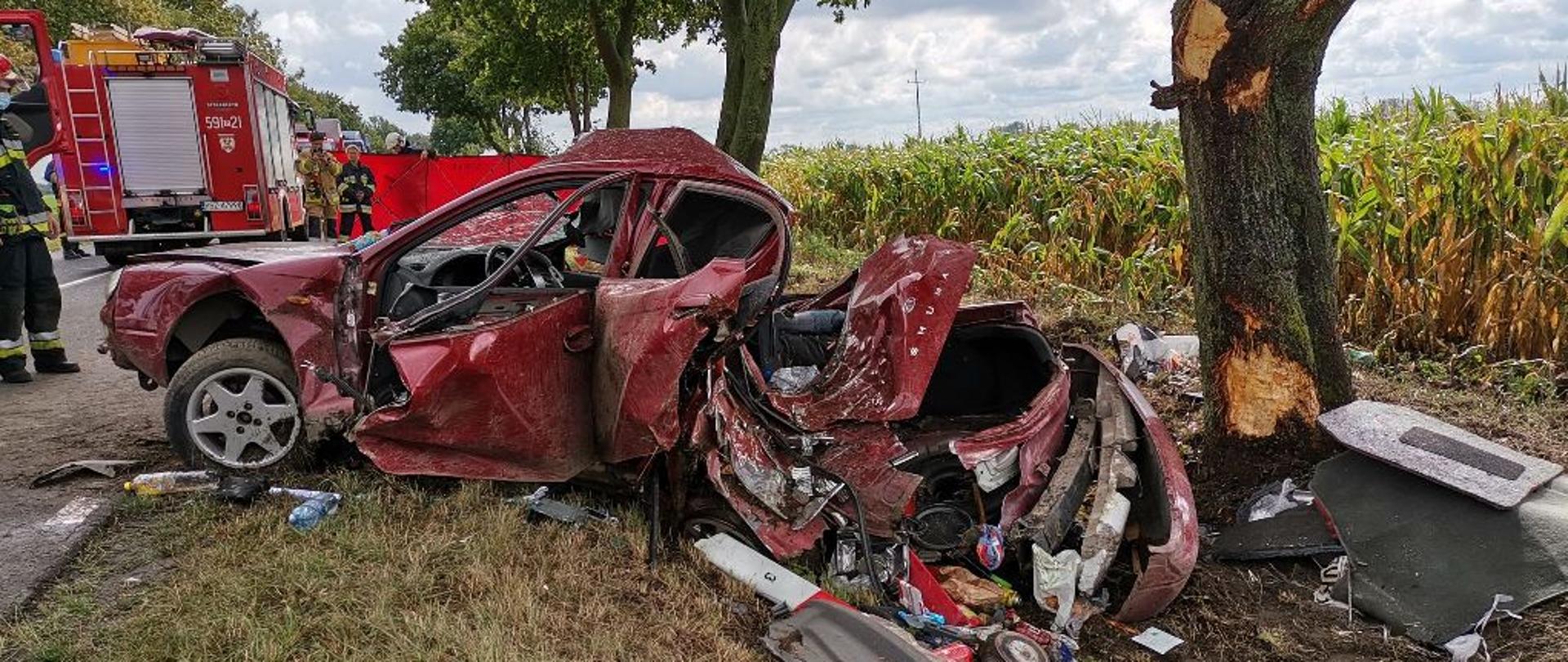 Samochód osobowy znajduję się w pobliżu drzewa, całkowicie uszkodzony, w oddali widać samochód straży pożarnej