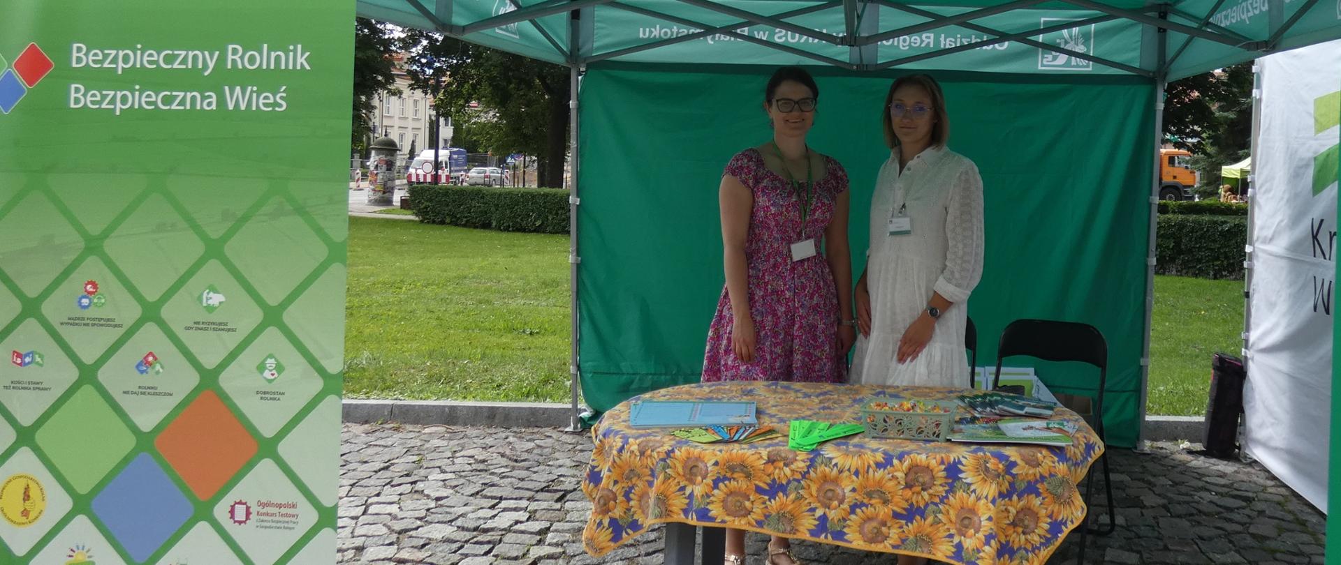 Plenerowe stoisko informacyjne Kasy Rolniczego Ubezpieczenia Społecznego. W centrum, pod dużym zielonym namiotem z białym napisem „Oddział Regionalny KRUS w Białymstoku”, stoją dwie uśmiechnięte kobiety – pracownice instytucji. Przed nimi stoi stolik nakryty obrusem w słoneczniki, na którym wyłożone są ulotki i broszury edukacyjne. Po lewej stronie namiotu ustawiony jest wysoki, zielony roll-upy. Baner zawiera hasło „Bezpieczny Rolnik, Bezpieczna wieś” oraz mozaikę logotypów i adres strony internetowej KRUS. Całość znajduje się na brukowanym placu, a w tle widać zielony trawnik i korony drzew.
