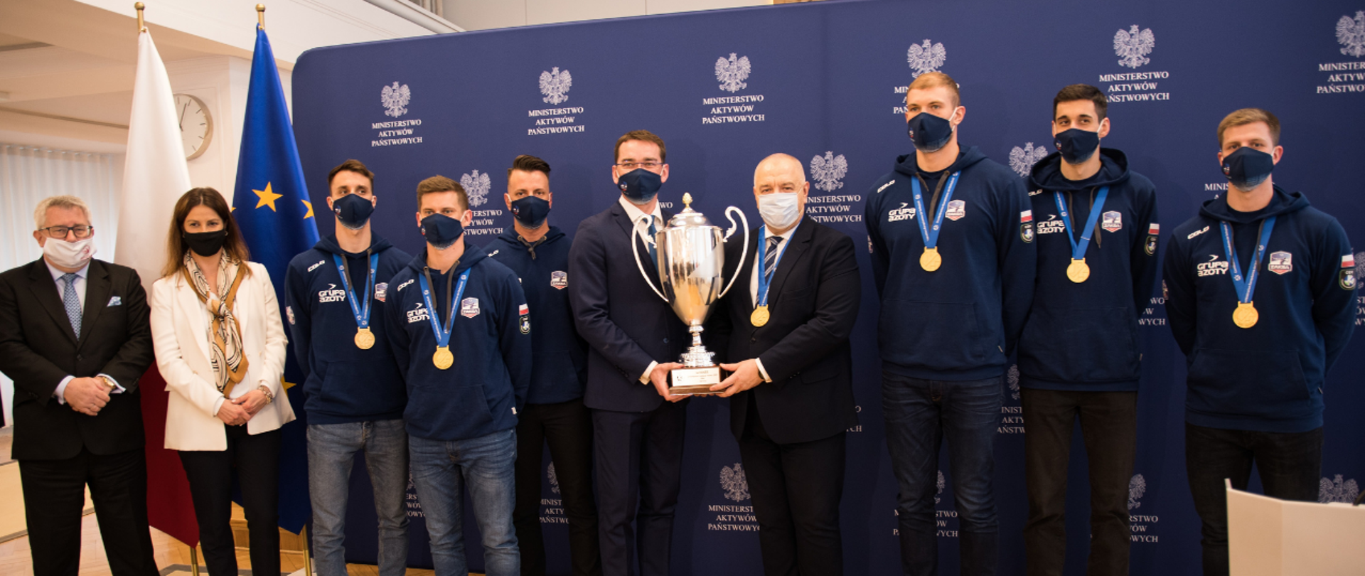 Na tle ścianki z logotypami MAP oraz flag polskiej i unijnej stoją w jednym rzędzie uczestnicy briefingu. Na środku wicepremier Jacek Sasin i prezes klubu Sebastian Świderski trzymają razem puchar zdobyty przez Zaksę.