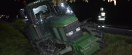 Wypadek na drodze wojewódzkiej nr 309. Zderzenie trzech pojazdów. Na pierwszym planie ciągnik rolniczy john deere koloru zielonego. Pojazd znajduje się w rowie. Obok na jezdni jest strażak w białym hełmie.