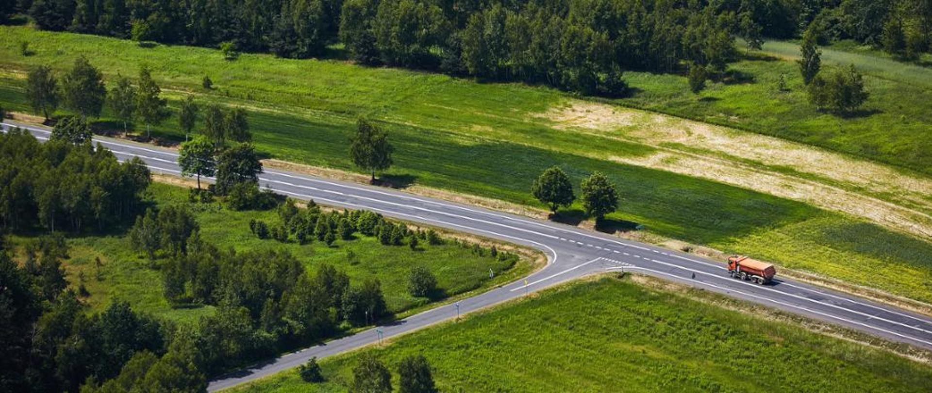Widok z lotu ptaka na dwupasową drogę biegnącą przez zielone, pagórkowate pola i łąki. Po obu stronach drogi rośnie wiele drzew i krzewów, a dalej widać gęsty las. Po drodze jedzie jedna ciężarówka, widoczna w dolnej części zdjęcia.