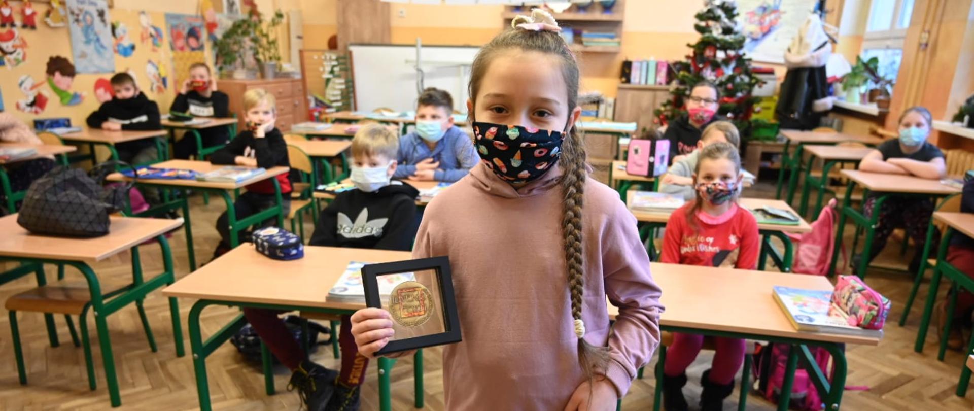 Na pierwszym planie dziewczyna, stoi w sali szkolnej na tle kolegów siedzących w ławkach. dziewczynka trzyma w ręku odznaczenie komendanta głównego PSP Luvenis Forti .