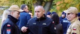 Zbliżenie na strażaka w czarnym polarze z nazwiskiem „Banaś Krzysztof” oraz gwiazdkami na piersi, który rozmawia i gestykuluje w stronę dwóch innych strażaków stojących po jego bokach. Mężczyzna po lewej nosi ciemną kurtkę i beżową czapkę z daszkiem, a strażak po prawej beżowy mundur i czapkę. W tle widać inne osoby, w tym funkcjonariuszy Policji.