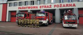 siedmiu strażaków ubranych w stroje specjalne znajduje się na tle trzech czerwonych samochodów ratowniczo gaśniczych za nimi znajduje się budynek PSP w Łowiczu