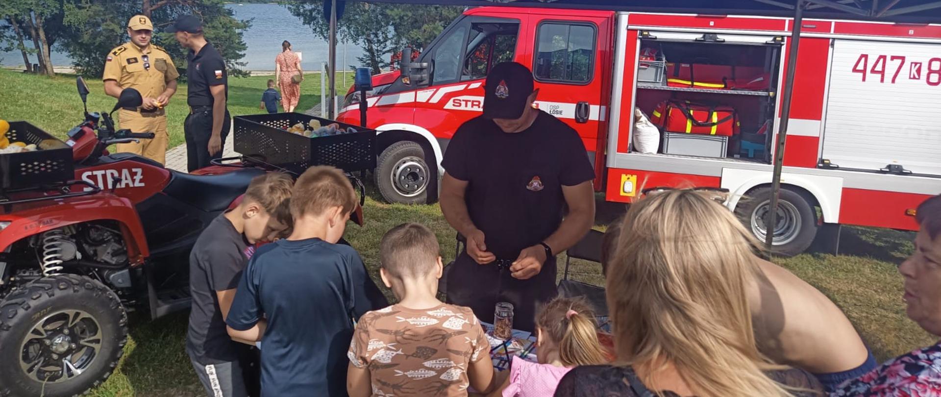 Na zdjęciu widoczna grupa dzieci i osób dorosłych uczestniczących w pikniku rodzinnym nad zalewem Klimkówka przed stanowiskiem wystawienniczym Komendy Powiatowej Państwowej Straży Pożarnej w Gorlicach. W którym strażacy prowadzili prelekcje i konkursy z zakresu bezpieczeństwa przeciwpożarowego i bezpieczeństwa nad wodą. W tle widoczne samochody pożarnicze koloru czerwonego.