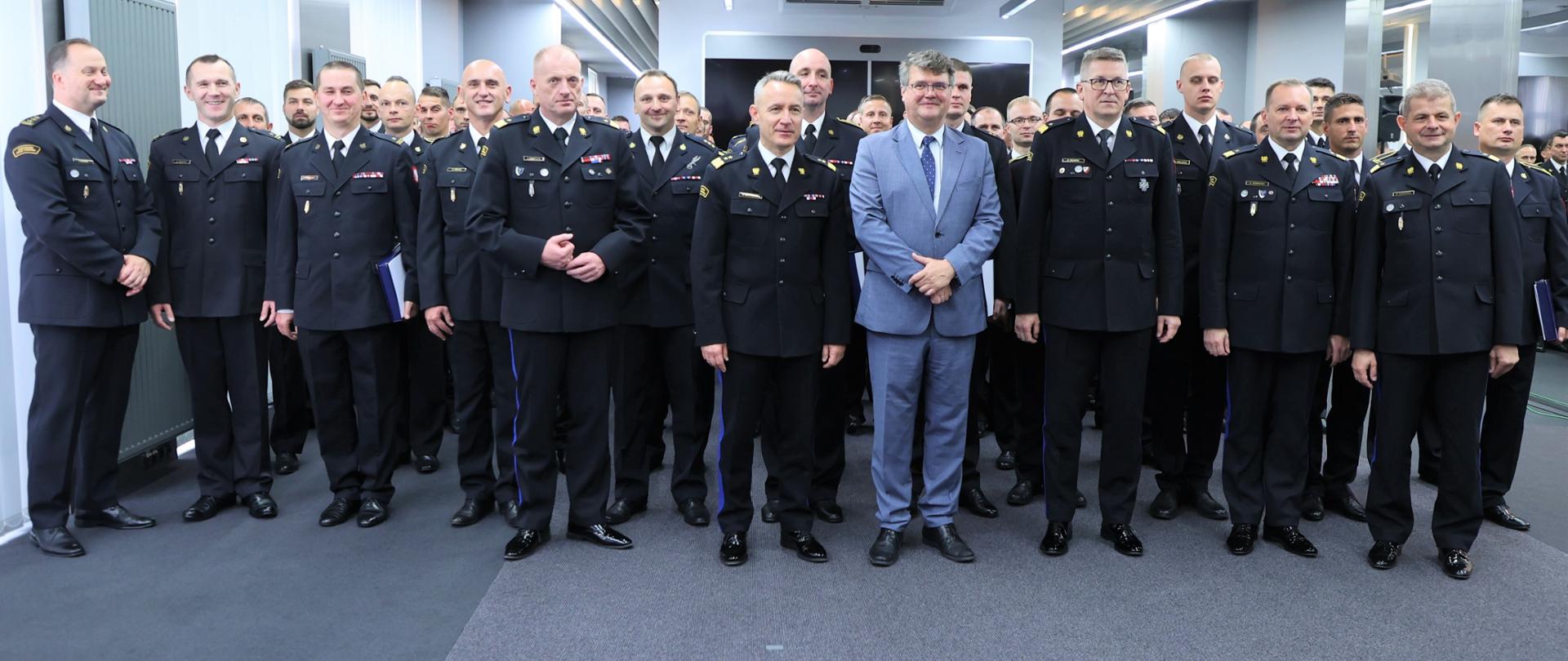 Na zdjęciu widać strażaków którzy brali udział w akcji gaszenia pożarów lasów we Francji w mundurach wyjściowych ustawionych obok siebie na sali konferencyjnej, oraz komendanta głównego PSP gen. brygadiera Andrzeja Bartkowiaka wraz z zastępcami, komendantami województw dolnośląskiego, zachodniopomorskiego i wiceministra SWiA Macieja Wąsika ustawionych razem do zdjęcia grupowego. 