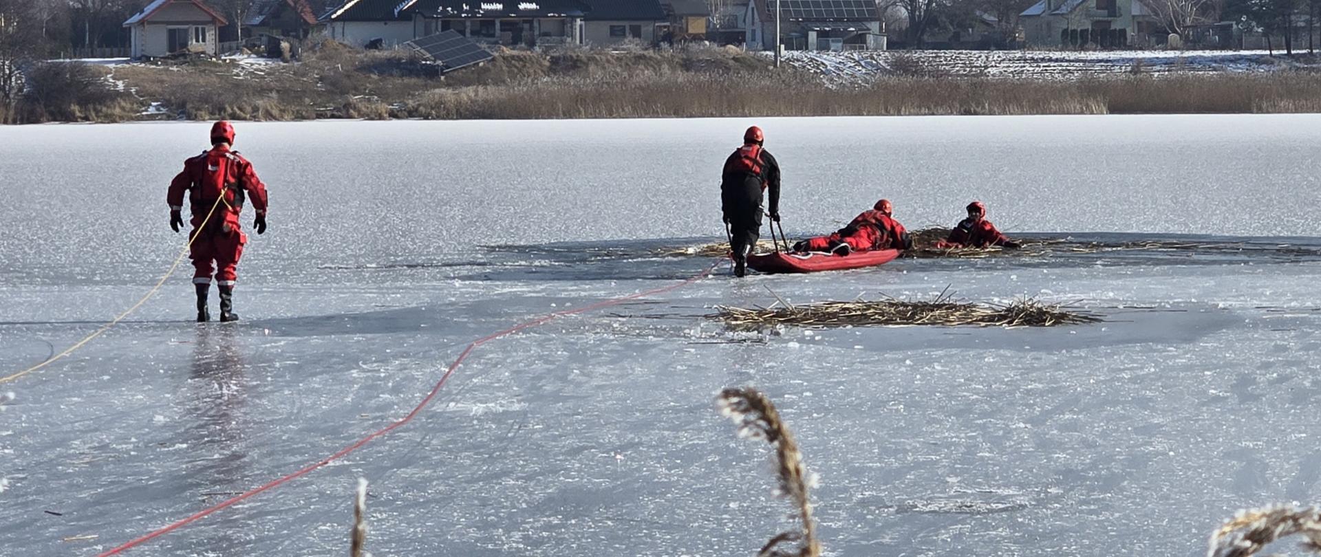 Zdjęcie przedstawia strażaków PSP ubranych w suche skafandry oraz kamizelki ratownicze, którzy poruszają się po zamarzniętym jeziorze. Do swoich kamizelek mają przypięte liny asekuracyjne. Przed nimi w przeręblu czeka strażak-pozorant.