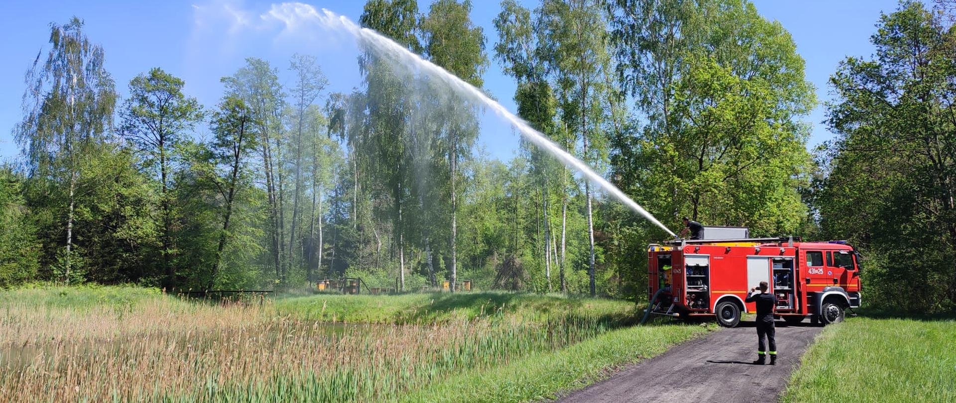Zastęp straży stoi na drodze w lesie i pobiera wodę. Następnie leje ją poprzez działko wodne.