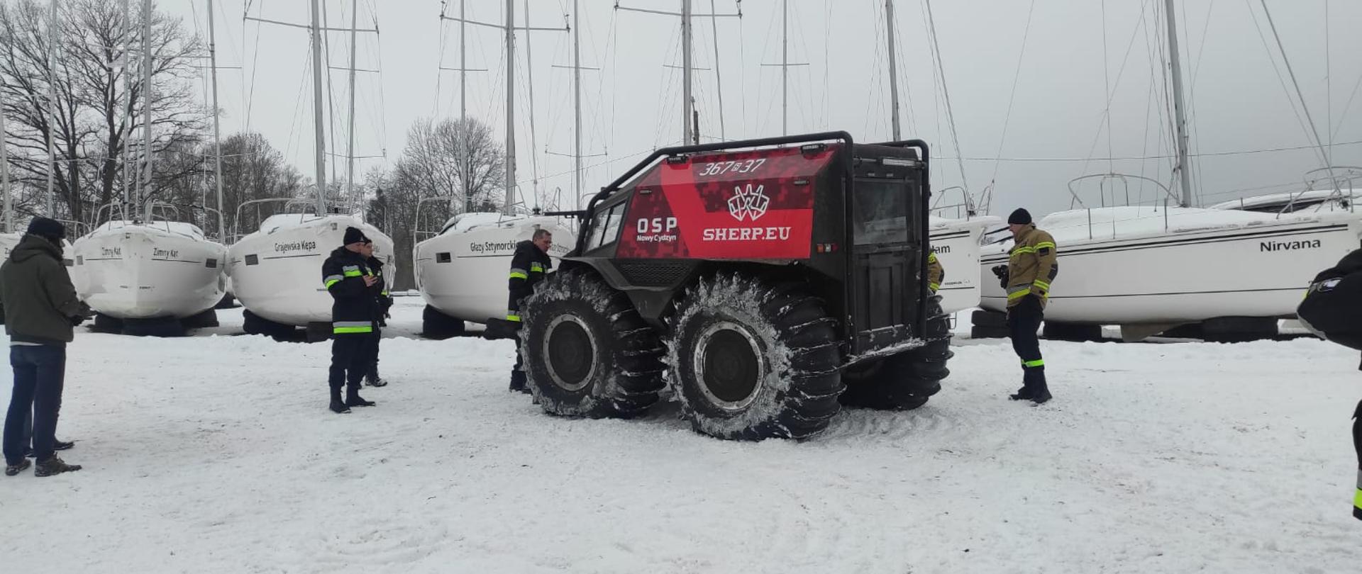 Na zaśnieżonym terenie obok jeziora stoi pojazd z wielkimi kołami. Obok strażacy. Z tyłu łodzie