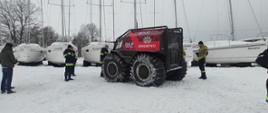 Na zaśnieżonym terenie obok jeziora stoi pojazd z wielkimi kołami. Obok strażacy. Z tyłu łodzie.