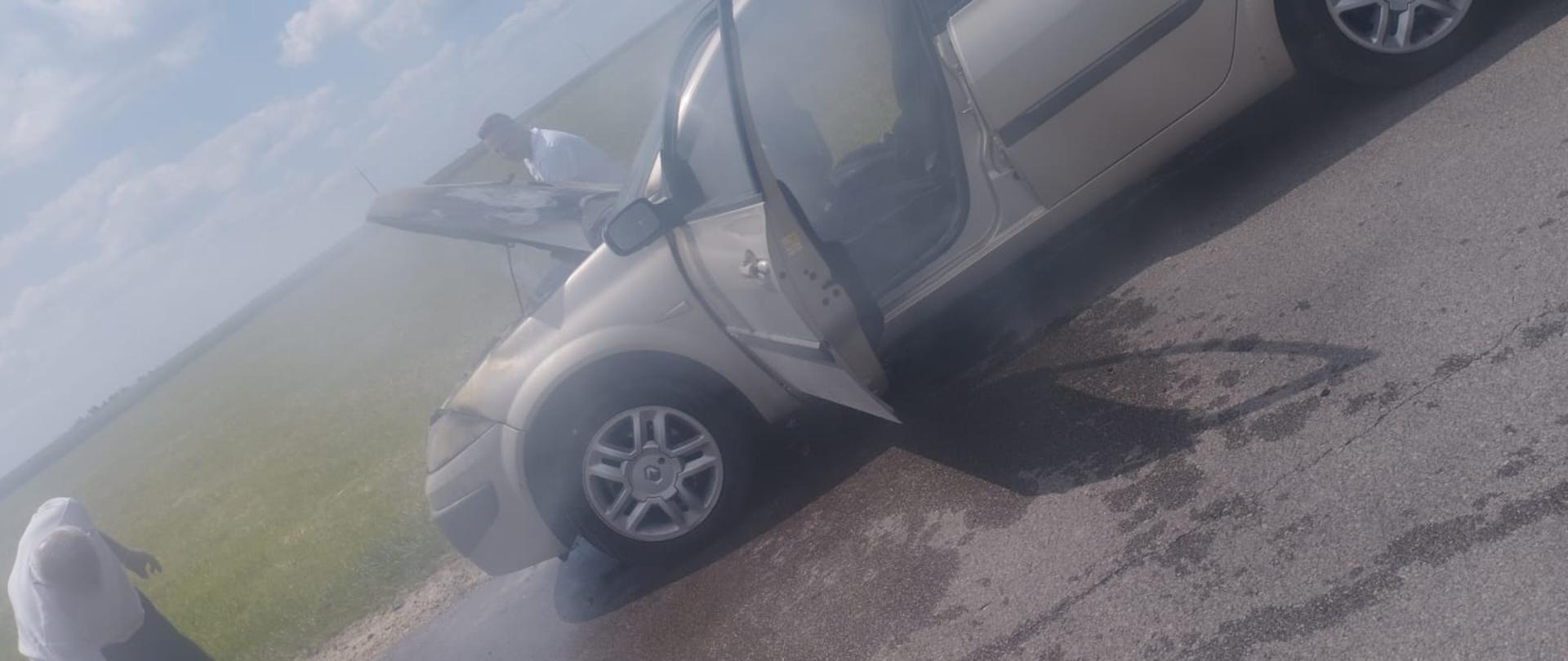 Na poboczu drogi stoi zadymiony szary samochód osobowy. Obok widać dwóch mężczyzn w białych podkoszulkach. W tle zielona trawa na łące.