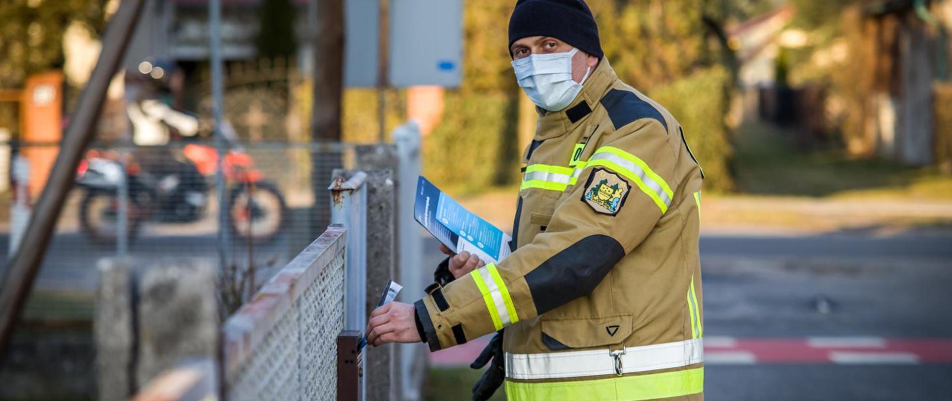 Zdjęcie przedstawia strażaka OSP wrzucającego ulotkę propagujące wiedzę na temat profilaktyki oraz szczepień przeciwko COVID-19. Strażak wrzuca ulotkę lewą ręką do skrzynki na listy. W prawej ręce trzyma pozostałe ulotki. Strażak ubrany w ubranie specjalne koloru piaskowego, czarną czapkę i jasną maseczkę na twarzy.