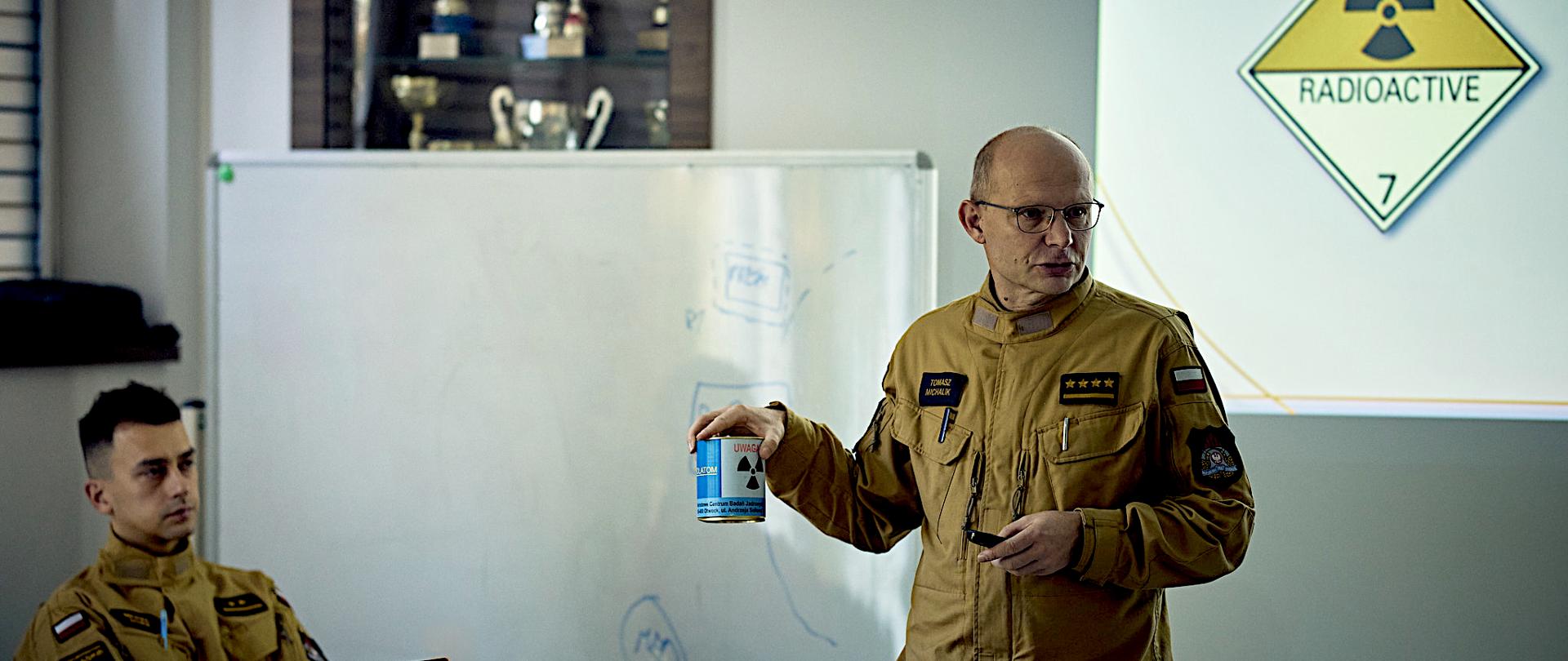 Zdjęcie przedstawia scenę ze specjalistycznego szkolenia mającego miejsce w typowej sali wykładowej. W centrum uwagi znajduje się strażak ubrany w jasny, piaskowy mundur służbowy, który prowadzi wykład. Mężczyzna ma okulary i w prawej dłoni prezentuje metalową puszkę z wyraźnym, niebiesko-żółtym ostrzeżeniem o radioaktywności. Za jego plecami, na ekranie, jaskrawo świeci międzynarodowy znak niebezpieczeństwa radiologicznego ("RADIOACTIVE 7"), który dominuje w prawej części kadru. Po lewej stronie, na tablicy, widać ledwie widoczne schematy i notatki. Przy stole, po lewej stronie, siedzi strażak w takim samym mundurze. Patrzy na prowadzącego. W gablocie na ścianie, stoją puchary.