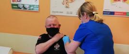 Zdjęcie przedstawia druha z jednostki Ochotniczej Straży Pożarnej (w umundurowaniu koszarowym) oraz pielęgniarkę podającą mu szczepionkę przeciwko COVID-19.