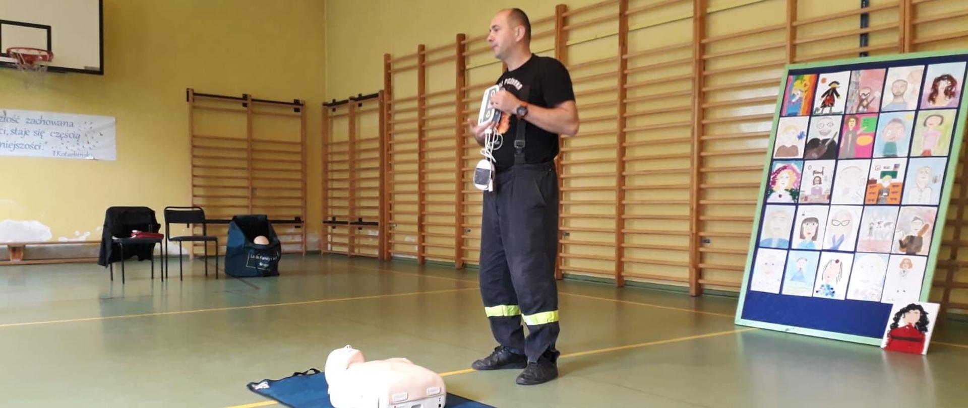 Zdjęcie przedstawia wnętrze sali gimnastycznej, gdzie na podłodze leży fantom medyczny. Za nim stoi strażak i prowadzi instruktaż użycia automatycznego defibrylatora.