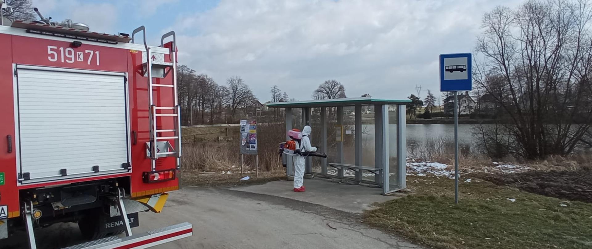 Strażak zabezpieczony w ubranie przeciwochlapaniowe dezynfekuje przystanek autobusowy. Po lewej stronie tył samochodu pożarniczego.
