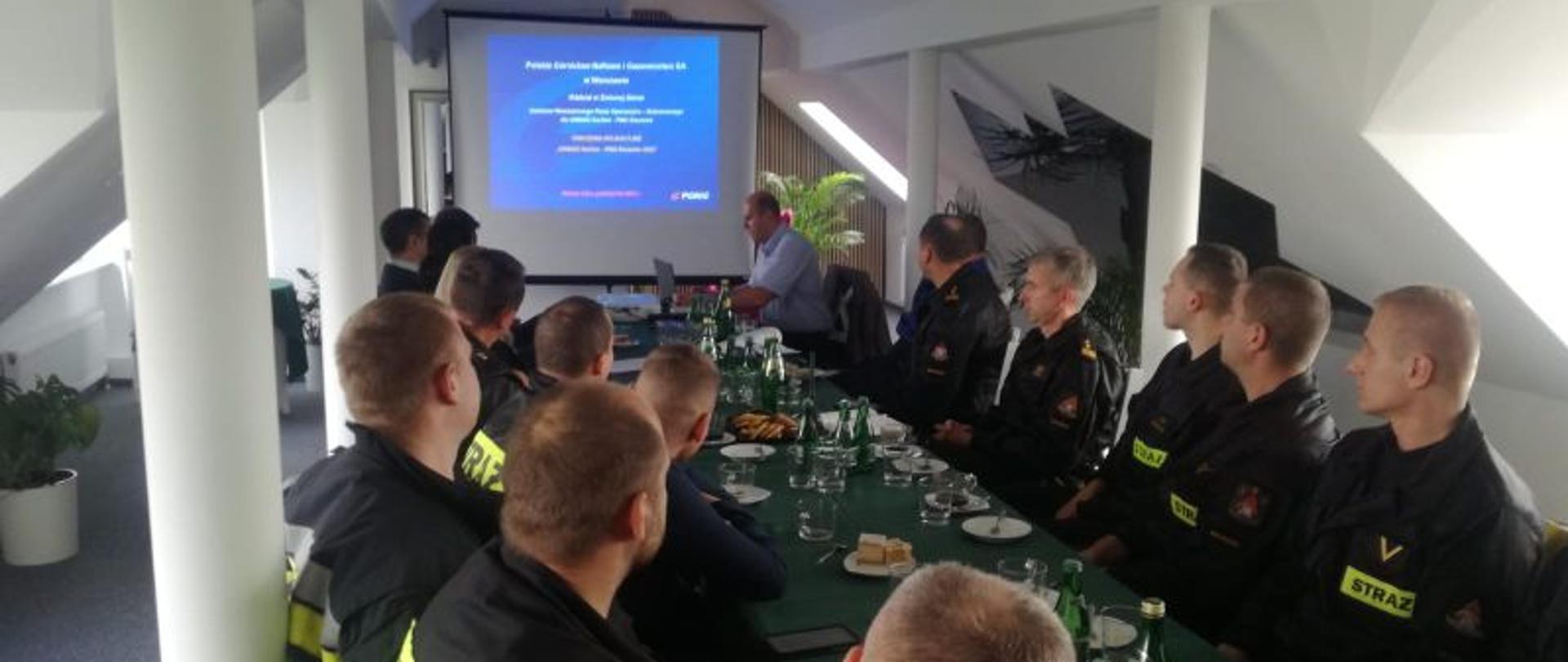 Zdjęcie wykonane wewnątrz budynku na sali szkoleniowej. Ściany białe, pomieszczenie niskie ze skośnym sufitem. Przy stole strażacy w ubraniach koszarowych. W oddali przy końcu stołu osoba prowadząca szkolenie.