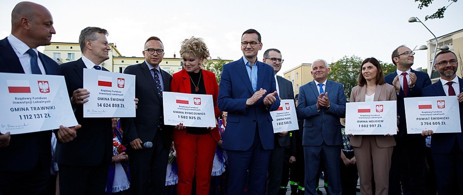 Premier stoi wśród ludzi trzymających czeki.