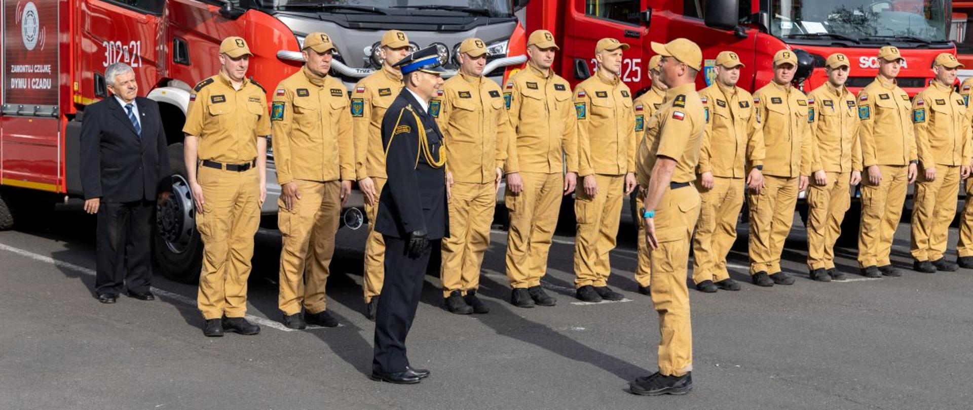 Grupa strażaków w jasnych mundurach stoi w zwartym szyku przed dwoma czerwonymi samochodami pożarniczymi. Przed szeregiem znajdują się dwie osoby w ciemnych mundurach galowych oraz mężczyzna w garniturze. Scena przedstawia oficjalny moment uroczystej zmiany służby w JRG 2 Opole.