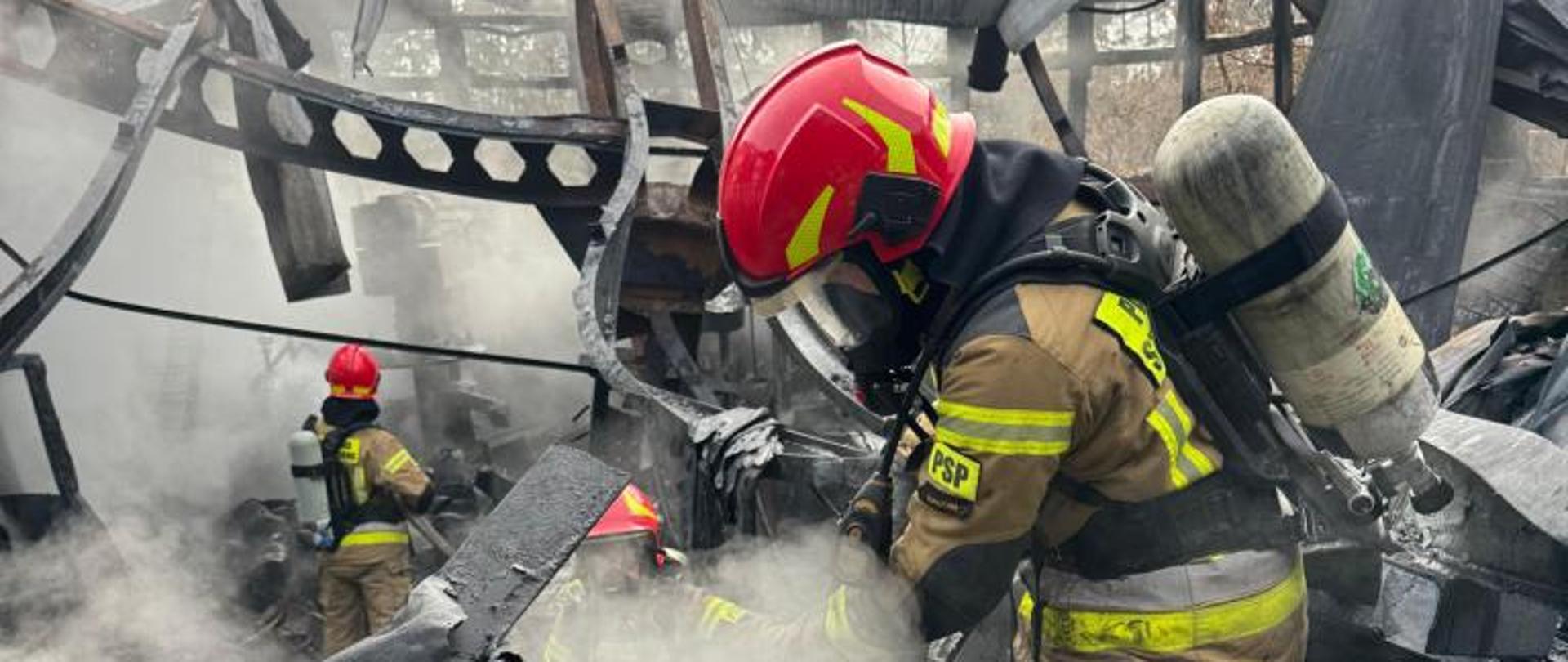Zdjęcie wykonane podczas akcji gaśniczej pożaru hali magazynowej w miejscowości Wierzbica. Na zdjęciu widać zawalony dach wraz z elementami konstrukcyjnymi hali. Pomiędzy nimi widać trzech strażaków dogaszających jeszcze palące się elementy wyposażenia. Strażacy ubrani w ubrania ochronne w kolorze piaskowym. Na głowach założone mają czerwone hełmy. Na twarzach posiadają maski ochronne chroniące przed trującymi substancjami znajdującymi się w dymi. Maska jest połączona przewodem z butlą z powietrzem znajdującą się na ich plecach. Ze spalonych resztek hali unosi się biały dym. 