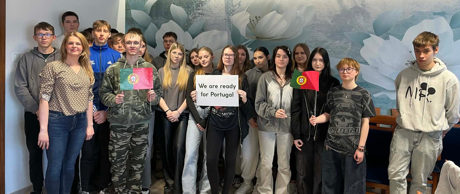 Grupa uczniów oraz nauczycielka pozują do zdjęcia, trzymając w rękach hasło "We are ready for Portugal" oraz portugalskie flagi z papieru.