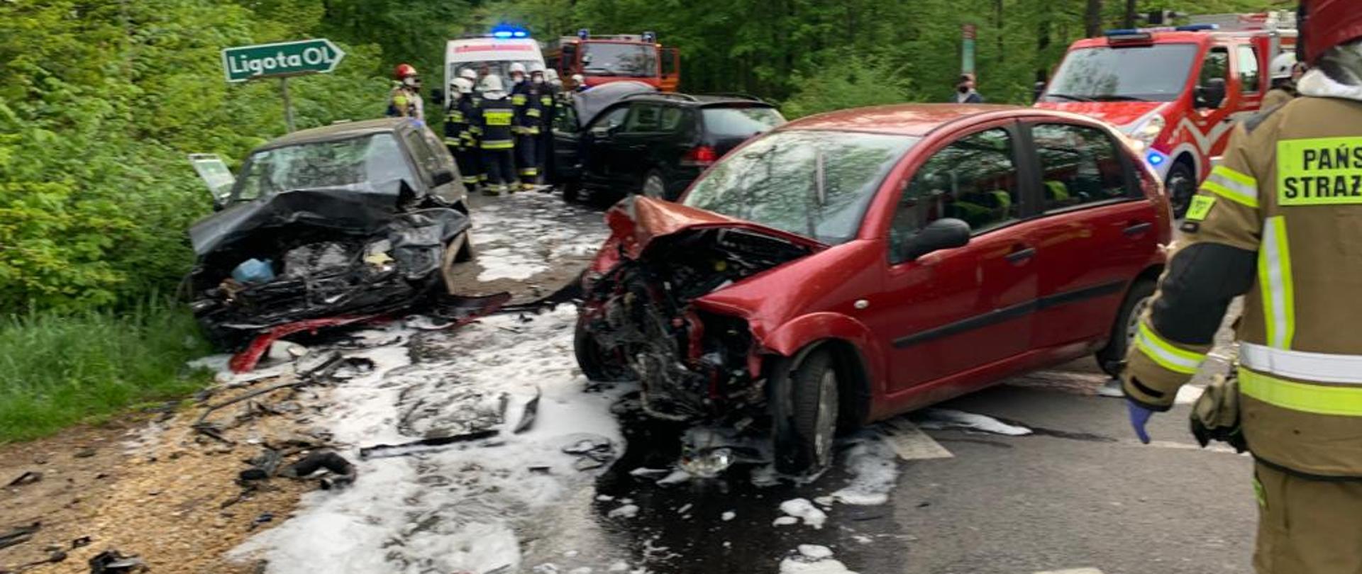 Na zdjęciu trzy samochody osobowe na drodze po zderzeniu, na drodze piana pożarnicza, samochody mają uszkodzone przednie części z tył widać samochody pożarnicze oraz karetkę pogotowia, w tle drzewa.