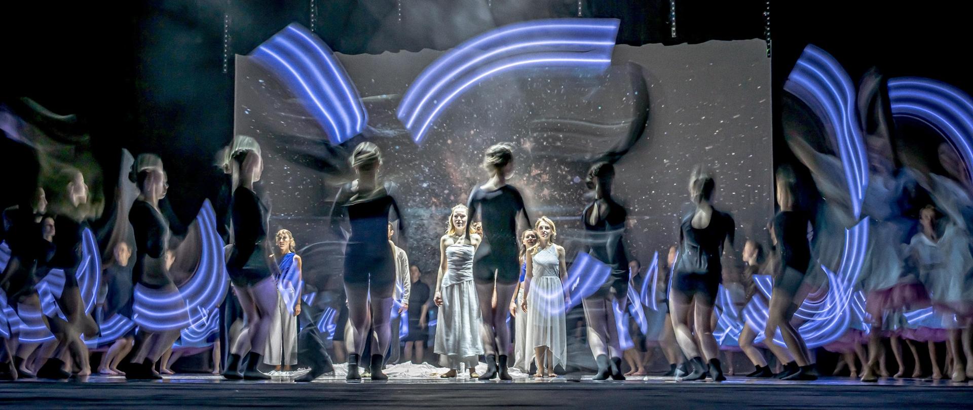Zdjęcie przedstawia grupę tancerek na scenie które stoją w układzie choreograficznym. Widoczne są jeszcze niebieskie smugi światła.