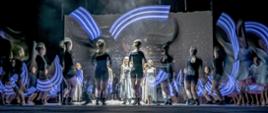 Zdjęcie przedstawia grupę tancerek na scenie które stoją w układzie choreograficznym. Widoczne są jeszcze niebieskie smugi światła.
